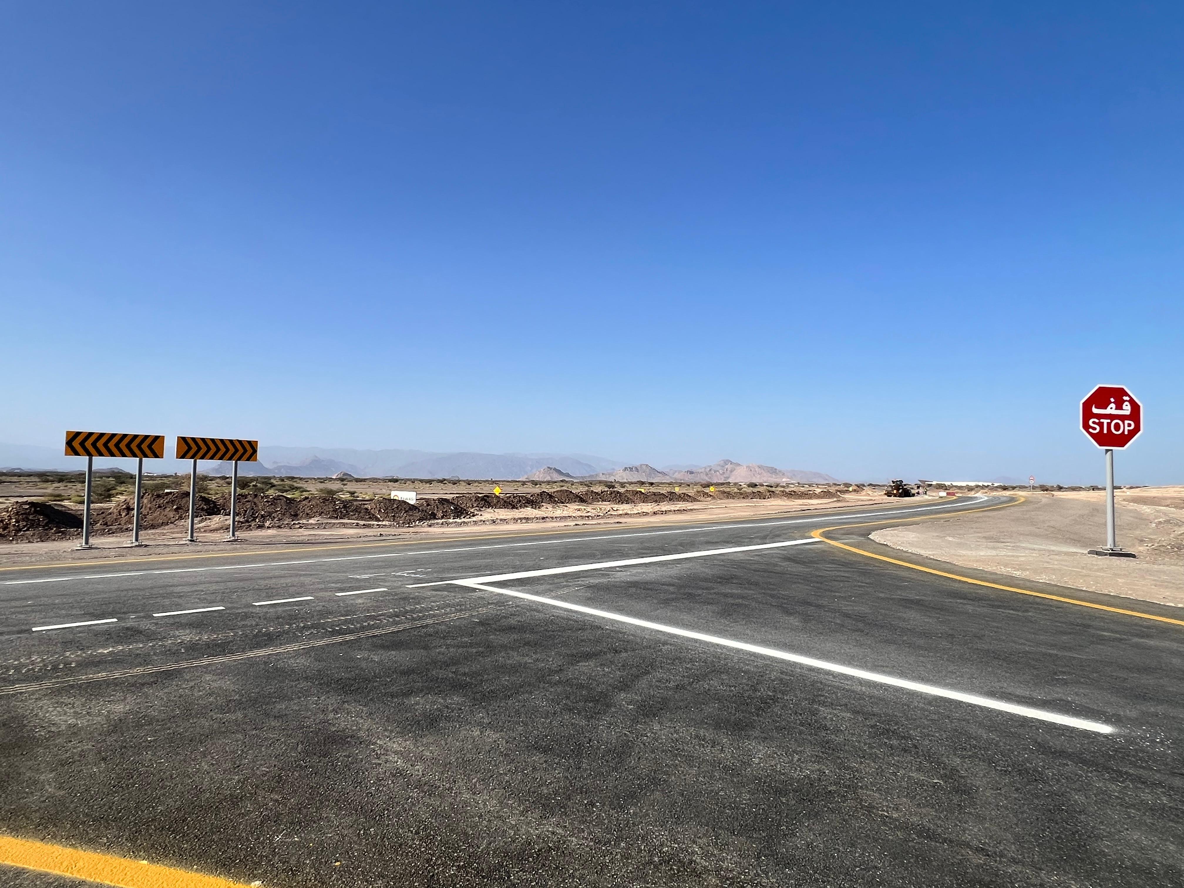 RRS-326 Oman Palm Development link road — finished road junction with chevron signage and mountain backdrop
